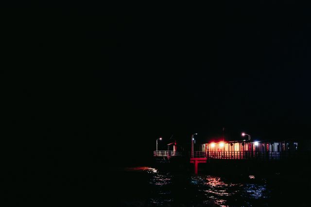 An illuminated pier stands out against the dark night, its lights reflecting off the tranquil seawater. Ideal for visuals in travel blogs, relaxation themes, or serene nightscape collections showcasing coastal or waterfront beauty.