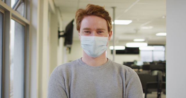 Young male professional standing in a modern office, practicing safety by wearing a protective mask. Ideal for topics related to workplace health, pandemic precautions, professional life, and office safety measures.