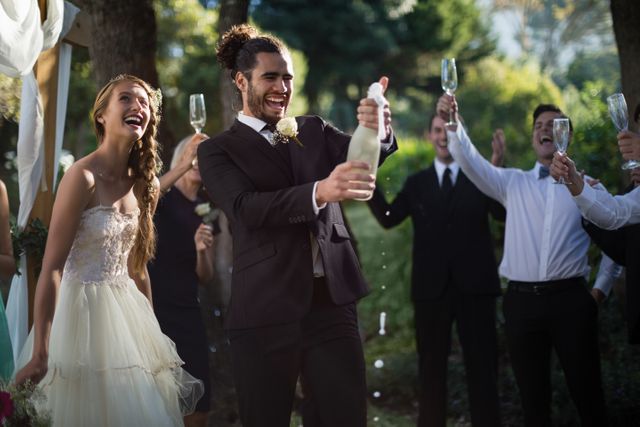 Groom opening champagne bottle surrounded by smiling bride and guests at outdoor wedding celebration. Ideal for use in wedding planning materials, celebration invitations, and articles about wedding traditions and joyful moments.