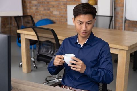 Professional Asian Man Viewing Monitor While Holding Coffee in Modern Office