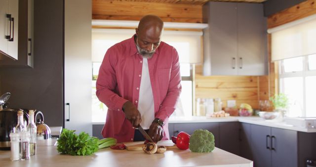 Senior man dressed casually and chopping vegetables, including broccoli and bell peppers, on kitchen counter. Modern kitchen with wooden cabinets and well-lit atmosphere, creating a sense of warmth and coziness. Ideal for use in lifestyle blogs, home cooking articles, or advertisements promoting healthy living and senior activities.
