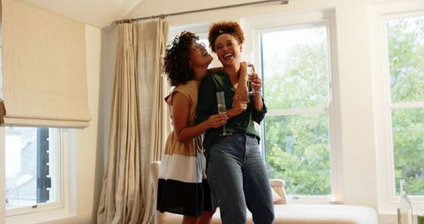 Two friends laughing and toasting with champagne flutes in sunlit cozy living room
