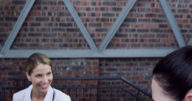 Foreground shows the back of a person's head with focus on a smiling Caucasian woman in the background, against a brick wall, with copy space. Her cheerful expression and casual business attire suggest a positive professional interaction or a friendly meeting outdoors.