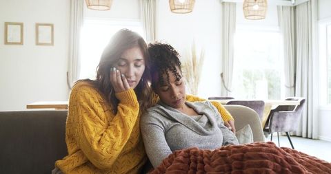 Two women sharing cozy blanket on sunlit living room sofa, comforting friendship moment