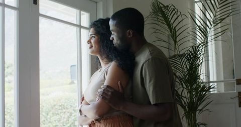 Sunlit diverse couple embracing by glass door, woman gazing outside, intimate home moment