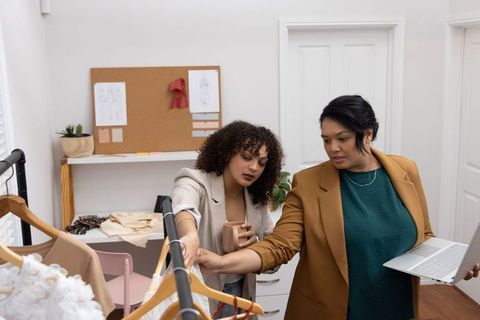 Multicultural women collaborating over garments on clothing rack in modern fashion studio