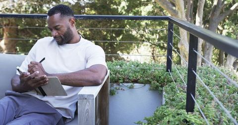 Man sitting on sunlit balcony writing in notebook, holding pen, relaxing among greenery