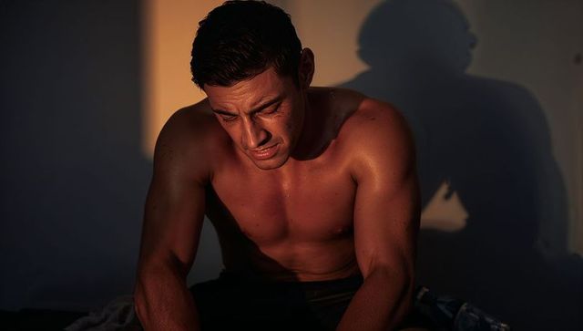 Shirtless athletic man sitting leaning forward sweating in moody low-light chiaroscuro