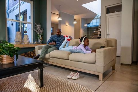 African american father and daughter relaxing on sofa using smartphone and tablet at home