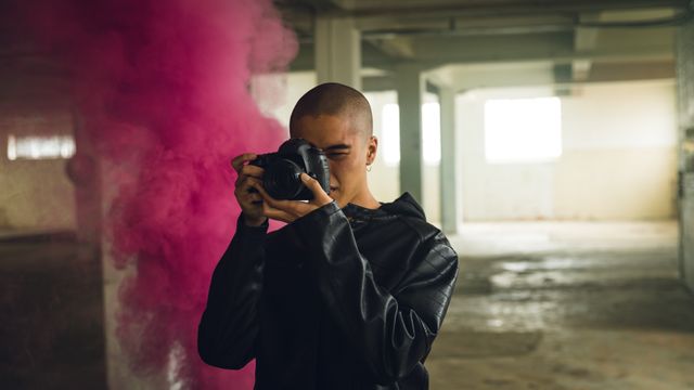 Front view of a hip young biracial man in an empty warehouse, taking photos with SLR camera, wearing black jacked, pink smoke bomb behind him.