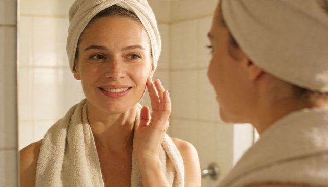 Smiling woman wearing towel and turban touching face at bathroom mirror for skincare