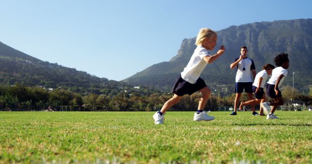Children dressed in sports attire running and playing on a green field during a sports day event, with mountains in the background and a coach observing. Suitable for education-themed projects, sports and outdoor activity promotions, and wellness advertising.