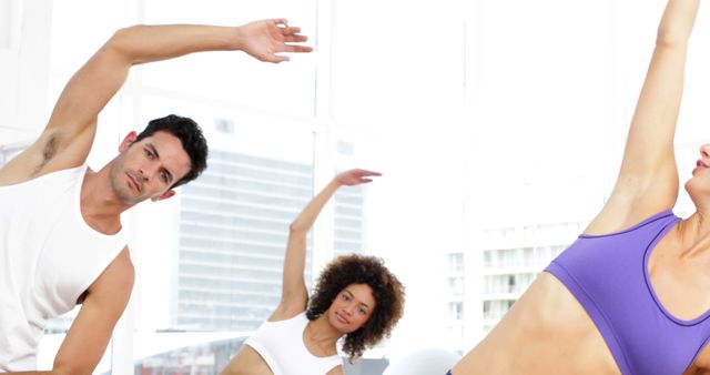 Group of diverse individuals performing stretching exercises in a modern gym with large windows. They are focused on flexibility and fitness. Ideal for advertisements and content related to fitness classes, healthy lifestyles, and group workouts.