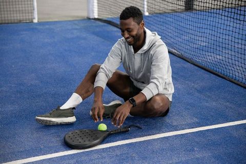 Smiling athlete reaching for yellow ball on blue paddle court while stretching in hoodie