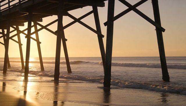 Golden-hour pier pilings casting geometric silhouettes over reflective wet sand and surf