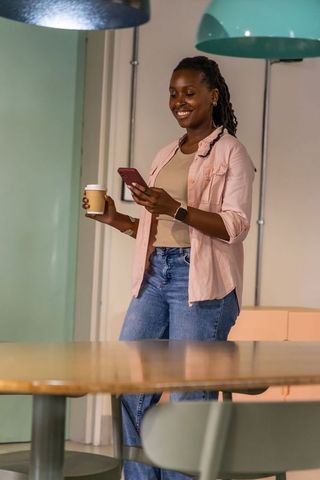 African american woman smiling and checking smartphone with coffee in modern coworking space