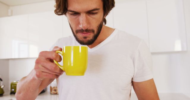 Focused caucasian man with beard holding cup of coffee and looking down, reading in kitchen. Morning, domestic life and lifestyle, unaltered.