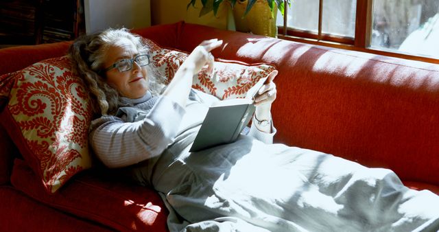 Senior woman comfortably reclining on a red sofa, reading a digital book with her glasses on. Warm sunlight illuminates her and the room features cozy decor with patterned pillows and houseplants. Picture can be used for illustrating lifestyles, leisure activities of elderly people, promote reading or digital book trends among seniors.
