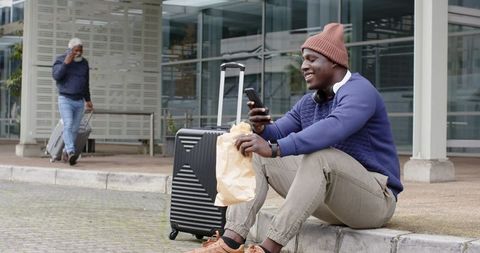 African american traveler sitting on curb checking phone beside rolling suitcase and snack
