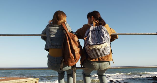 Two young women are sitting on a railing by the seaside with backpacks, enjoying the ocean view. The scene suggests friendship, adventure, and relaxation. Perfect for themes related to travel, leisure, friendship, nature, and oceanic landscapes. Ideal for use in travel websites, social media posts, and promotional materials for coastal destinations.