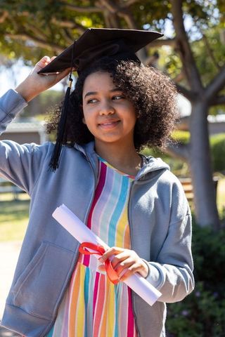 African american teen celebrating graduation wearing mortarboard and holding diploma outdoors