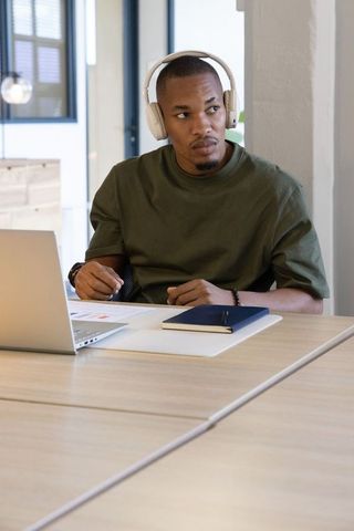 African american man wearing headphones working on laptop in coworking space with notebook