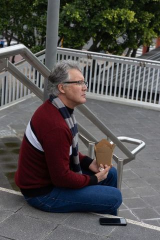 Senior man sitting on concrete steps in urban plaza holding takeout box and smartphone