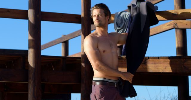 Man Holding Wetsuit Near Wooden Beach Structure on Sunny Day - Download Free Stock Images Pikwizard.com