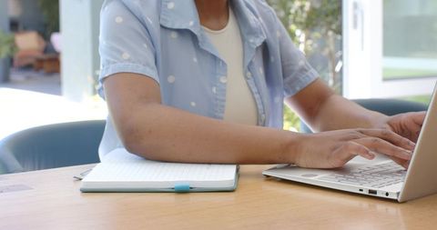 African teen female typing on laptop at home desk with notebook and natural light