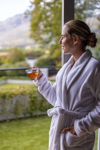Woman in white robe enjoying morning tea while gazing at mountain view from terrace