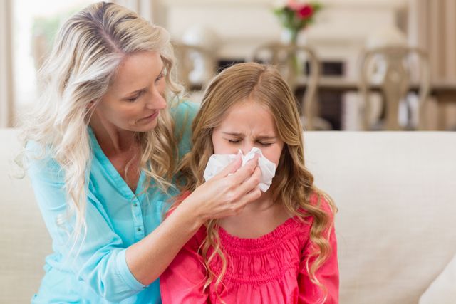 Mother helping daughter blowing her nose on sofa in living room at home