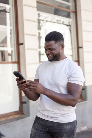 Smiling african american man checking smartphone on urban sidewalk in white tee
