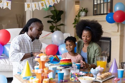 African american family celebrating birthday cutting rainbow cake with balloons and cupcakes