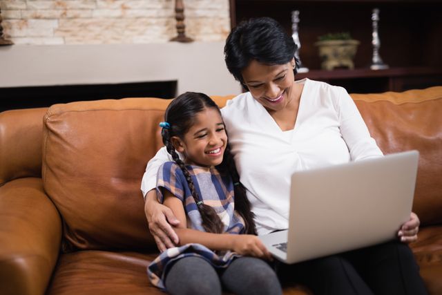 This image shows a woman and her granddaughter sitting on a sofa at home, smiling and using a laptop together. It is perfect for illustrating concepts of family bonding, intergenerational relationships, and the use of technology in everyday life. Ideal for use in articles, advertisements, or websites focused on family, education, technology, or home life.