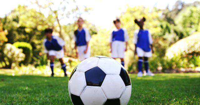 A soccer ball is in focus in the foreground, with a diverse group of young girls in sports uniforms blurred in the background, ready to play. Capturing the anticipation of a youth soccer game, the image emphasizes the importance of teamwork and physical activity among children.