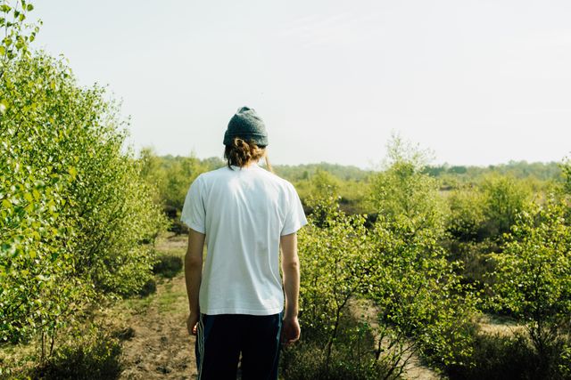 Young individual wearing beanie and white t-shirt walking through dense, green forest path. Ideal for themes of exploration, nature walks, solitude, personal adventure, outdoor activities, environmental consciousness, and travel.