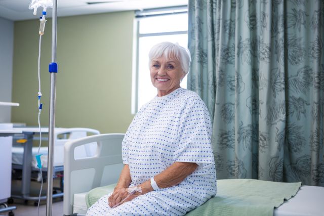Smiling senior woman sitting on hospital bed wearing a medical gown. She is receiving treatment with an IV drip attached to her arm. Suitable for use in healthcare advertising, senior care publications, and medical facility brochures.