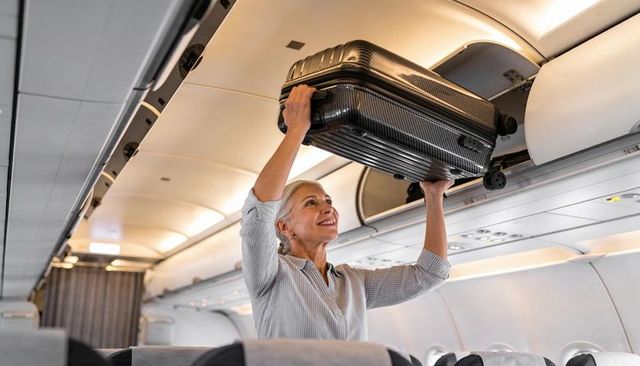 Senior woman placing carry-on into overhead bin inside airplane cabin smiling confidently