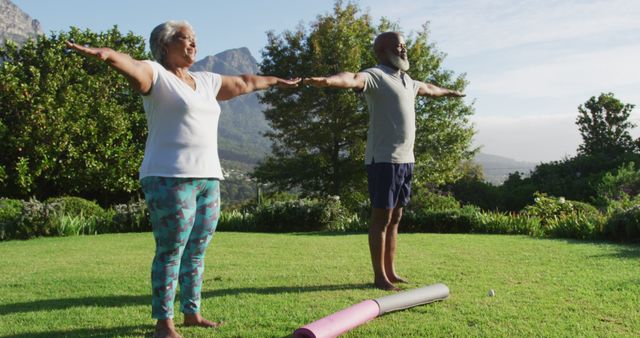 Senior couple engages in morning yoga routine in outdoor setting surrounded by mountains and trees. They are standing on grass, stretching arms wide, embracing the tranquility of nature. Health, wellness, and fitness concepts can utilize this image to promote exercise, outdoor activities, and a healthy lifestyle.