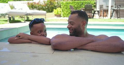 African american father and son relaxing at pool edge, smiling and chatting in backyard
