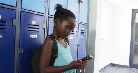 Teen student leaning by blue lockers checking smartphone in school hallway with backpack