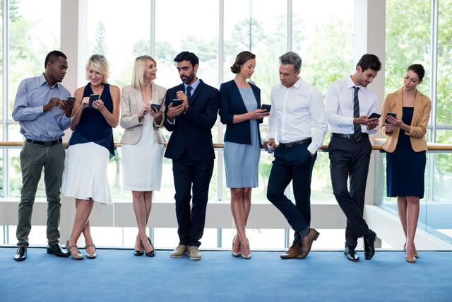 Group of business executives standing in a row, using mobile phones at a conference center. They are dressed in professional attire, indicating a formal business environment. This image can be used for themes related to business communication, networking, corporate events, teamwork, and the use of technology in professional settings.