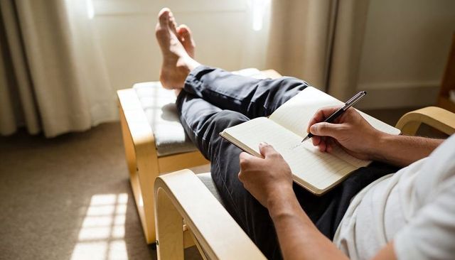 Barefoot man journaling on armchair in sunlit minimalist room, relaxed home writing scene