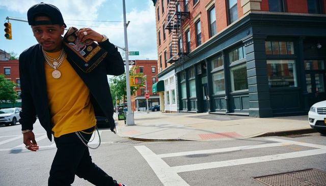 Streetwear man striding crosswalk wearing cap, gold chains, carrying patterned bag