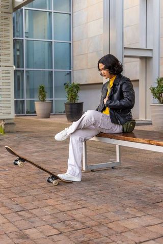 Urban skate style young woman sitting on bench with skateboard, leather jacket, yellow top
