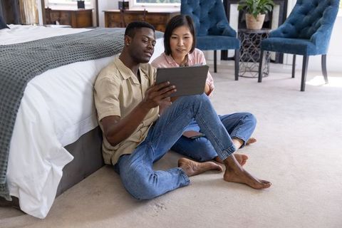 Interracial couple sitting on bedroom floor using tablet, planning homebuying together