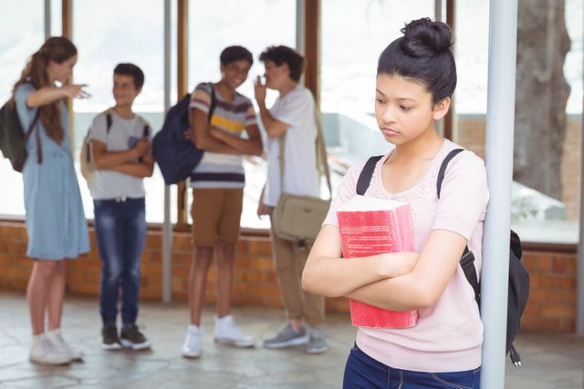 Teenage girl standing alone in a school corridor, holding books and looking sad while a group of students in the background are pointing and whispering. This image can be used to illustrate topics related to bullying, peer pressure, mental health issues among teenagers, and the importance of creating a supportive school environment.