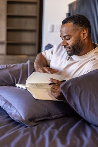 African american man journaling on bed lounging with notebook and pen in cozy sunlit room