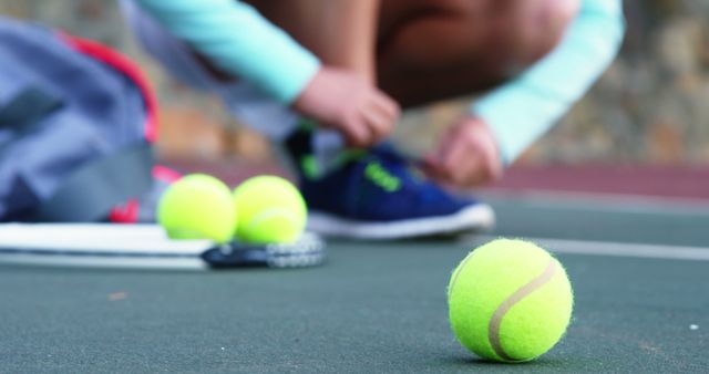 A close-up capturing a tennis player tying their sneakers on an outdoor court. Nearby, there are tennis balls and a racket on the ground. This image can be used for illustrating sports preparation, fitness, and active lifestyle themes on blogs, ads, and sports magazines.