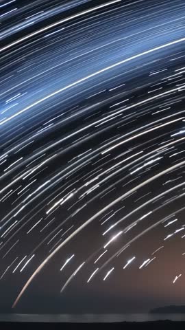 Vertical night timelapse capturing curved star trails over coastal horizon and tranquil sea